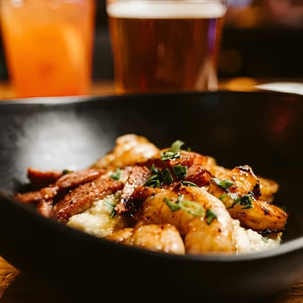 Shrimp and grits with bacon and chives in a black bowl, with drinks blurred in the background.