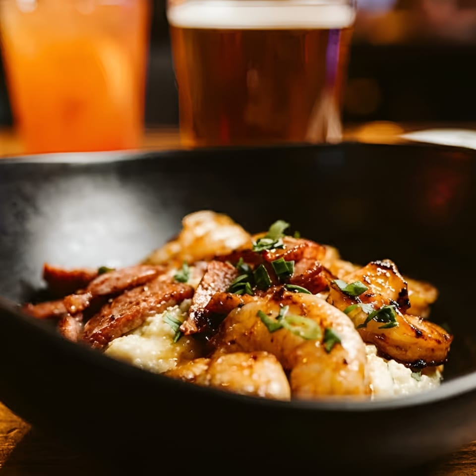 Shrimp and grits with bacon and chives in a black bowl, with drinks blurred in the background.