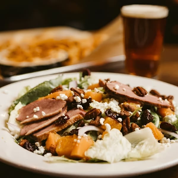 Duck breast salad with butternut squash, pecans, goat cheese, and dried fruit, with a beer in the background.