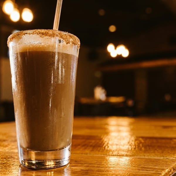 A tall glass of chocolate milkshake with a sugared rim and straw sits on a wooden bar.