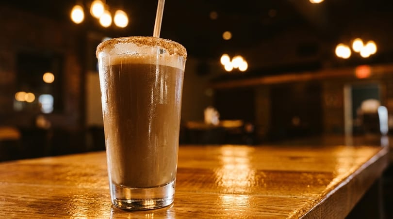 A tall glass of chocolate milkshake with a sugared rim and straw sits on a wooden bar.