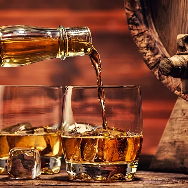 Whiskey pouring from a bottle into a glass with ice, next to another glass and a barrel.