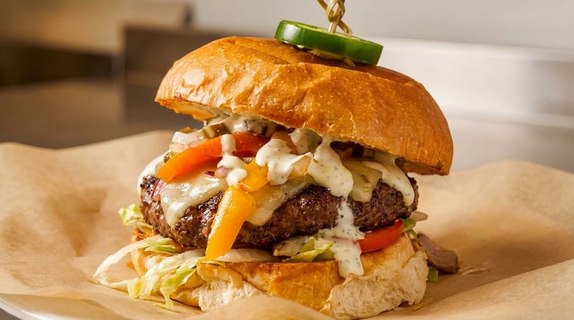 A juicy burger with melted cheese, colorful peppers, and creamy sauce, topped with a jalapeño slice.