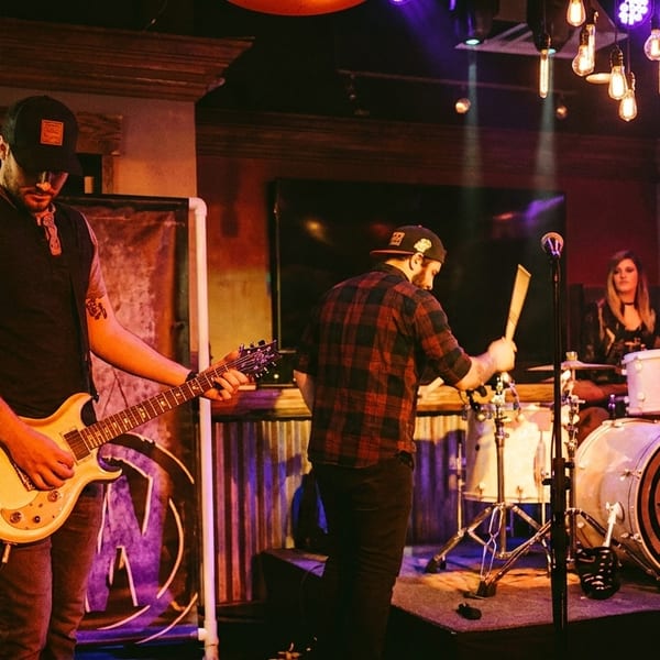 A band performs on stage with a guitarist, drummer, and female vocalist under purple lights.
