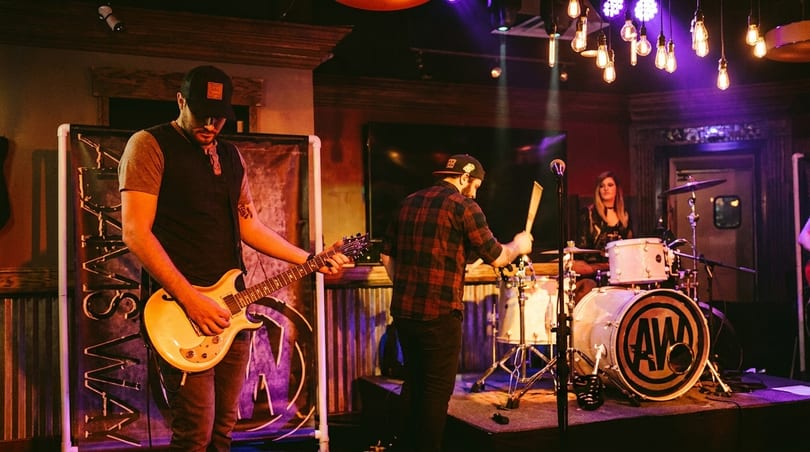 A band performs on stage with a guitarist, drummer, and female vocalist under purple lights.
